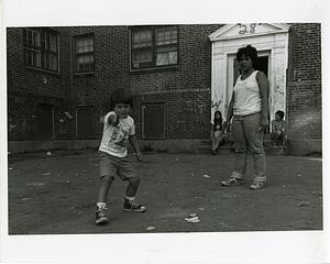 A young boy in a courtyard points at the camera while a teenager watches