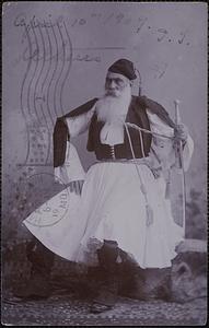 A bearded man sits wearing traditional Greek dress including a fustanella