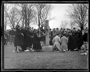 A crowd watches a group of women kneeling ands standing at a large stone crucifix, possibly a headstone