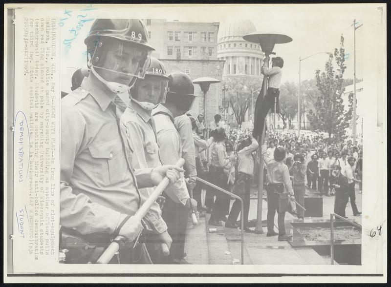 Down with Flag-- As long line of riot-equipment Madison, Wis., police guard city-county building as another officer pulls down red flag placed on pole marching University of Wisconsin students (background) today. Students are continuing their anti-police demonstration for third day. State capitol building is in background.