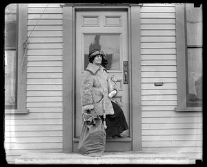 A woman stands by a door holding wrapped packages and papers, a sack by her side