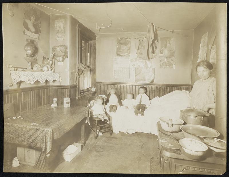 A woman and four children sit in their apartment, facing the camera. The room contains a stove, single bed, kitchen table, and laundry.