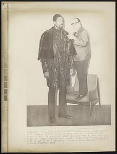 Fur Heaven's Sake, Bill--Player-coach Bill Russell of the Boston Celtics gets his 6 foot 11 inch frame fitted for a Persian lamb cavalier coat by New York fur designer Irving Hochberg who needs a chair for the fitting. The coat is capped by a detachable Alaskan seal cape.