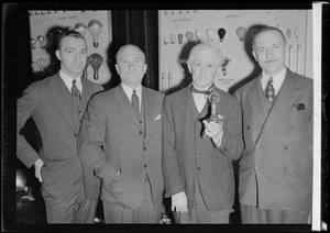 Four men standing before a light bulb display, one dressed like Thomas Edison