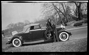 A woman sits on car hood holding a hat while posing with a man