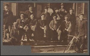 Group of men, most elderly and bearded, wearing hats, seated and standing on porch outside commercial establishment