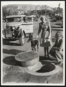 At Last - Wooden Manhole Covers - Wooden manhole covers popped up (or did they pop down?) in Los Angeles County where they were urgently needed to complete sewage facilities for a large war housing development. Developed by the county surveyor's office when customary cast-iron covers were not forthcoming because of the metal shortage, the new style street lids are reported to be decay-proof and termite-proof. They save 500 pounds of metal a piece, 250 pounds in the cover and 250 pounds in the frame. The covers come in two shapes--circular and hexagonal. This photo shows County Surveyor Alfred Jones explaining good points of wooden cover to Miss Beverly Hoyt. Big Black dog, "trina," seems bored.