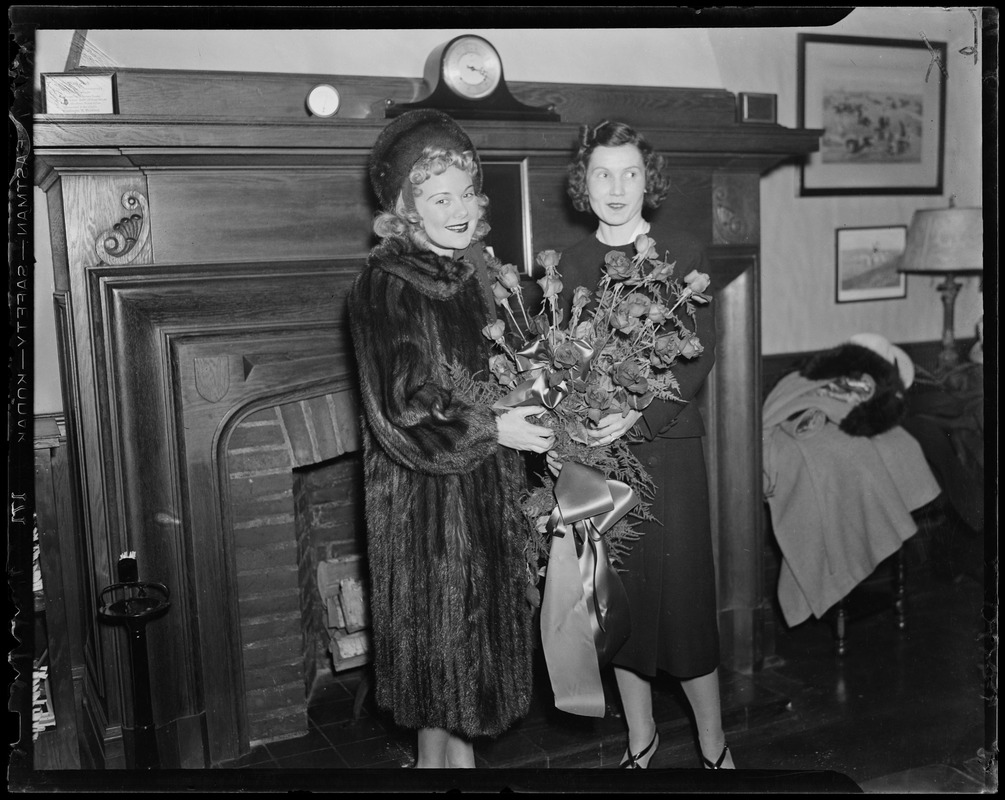 Sonja Henie holding flowers and posing with another woman in front of a fireplace
