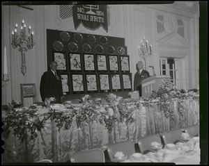 Two men standing by podium with wall featuring the athletes honored at the seventh B'nai B'rith sports dinner at the Sheraton Plaza