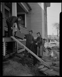 Man handing another man a suitcase from the porch of a damaged house