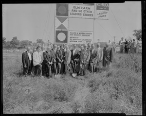 Group of men, some with shovels, posing in front of "The site of New England Shopping Center" sign
