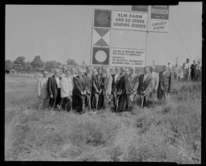 Group of men, some with shovels, posing in front of "The Site of New England Shopping Center" sign