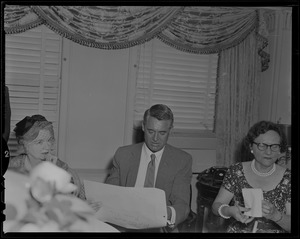 Cary Grant seated at a table in between two women and pulling something out of an envelope