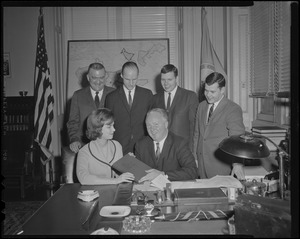 Mayor Collins seated at a table with a woman holding The Book of Mormon as four men standing look on