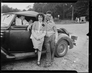 A man and woman lean against a car while two children look on from inside of the car