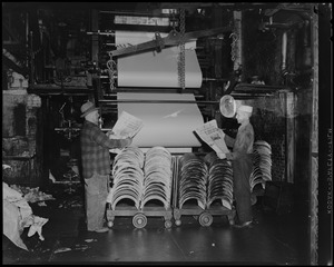Two men holding newspapers next to printing machines