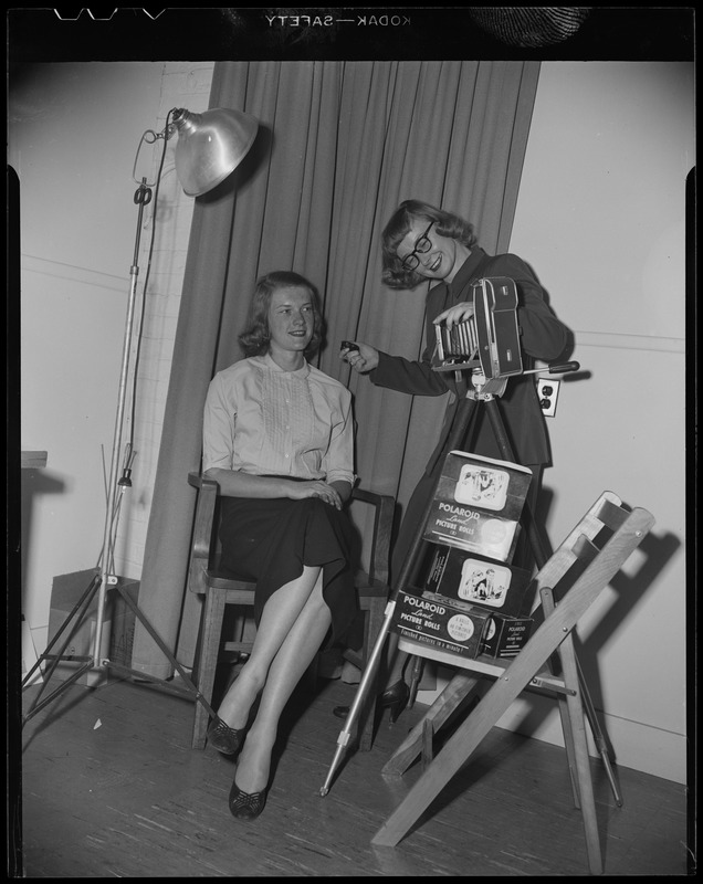 A woman poses in front of a Polaroid camera as another woman takes the photo