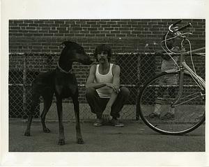 A man squats in a courtyard next to a dog and a bicycle