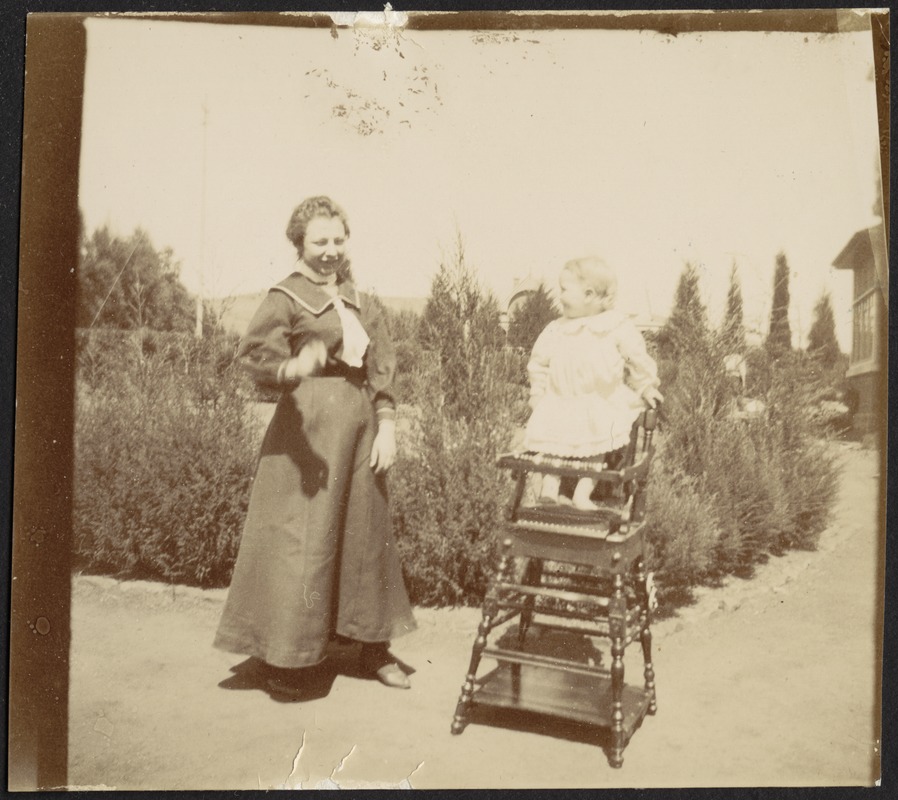 Woman standing next to small child in wooden high chair in garden