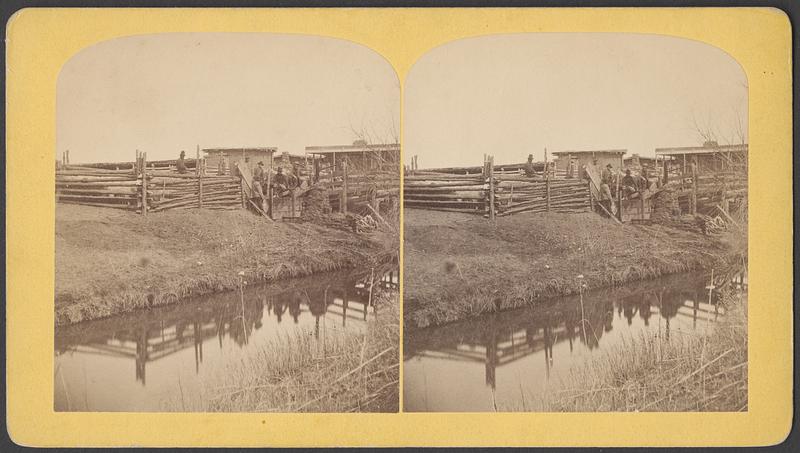 A group of men sit by a pen full of sheep, water in the foreground