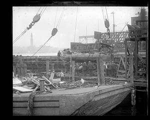 A crowd looks at an unidentified, possibly damaged item in the background, while there is a flat bottomed boat with debris on top of it in the foreground