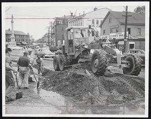 After the Flood comes cleanup. A road grader piles up dirt on Lowell St., across from City Hall, after rushing waters in Peabody square subsided yesterday. Then began cellar pumping-out operations and more water in streets. Officials said a record 5.74 inch rainfall was responsible for the high water.