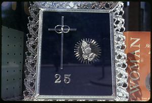 Framed item showing a cross with two interlinked rings, a pair of hands in prayer and the number 25