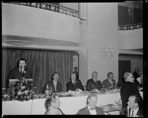 Man at the Statler Hotel podium speaking with panel table at his side
