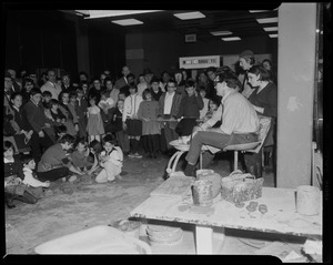 Man on a pottery wheel, demonstrating the process to a crowd, with several children working with clay on the floor in front of him