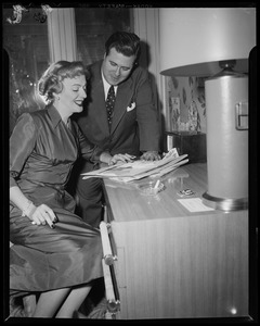 Christine Jorgenson sitting at a desk looking at a newspaper with a man by her side