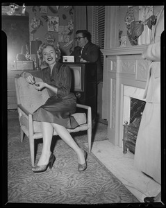 Christine Jorgenson seated by a fireplace with a man in the background
