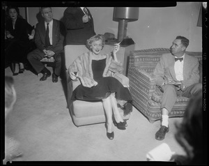 Christine Jorgenson seated, smoking and wearing a fur shawl, among several men and one woman