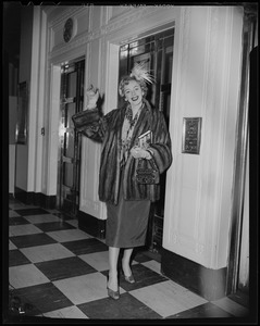 Christine Jorgenson waving in a fur coat, in front of elevators