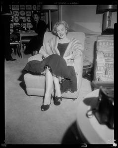 Christine Jorgenson sitting in chair, wearing fur coat, with woman in background
