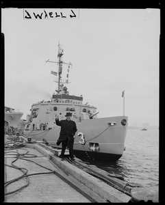 Man waving while standing on the dock by the bow of the U.S.S. Atka