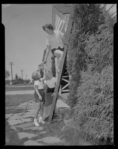 Girl passing a hammer to a woman on a ladder while another girl holds roof shingles beside them