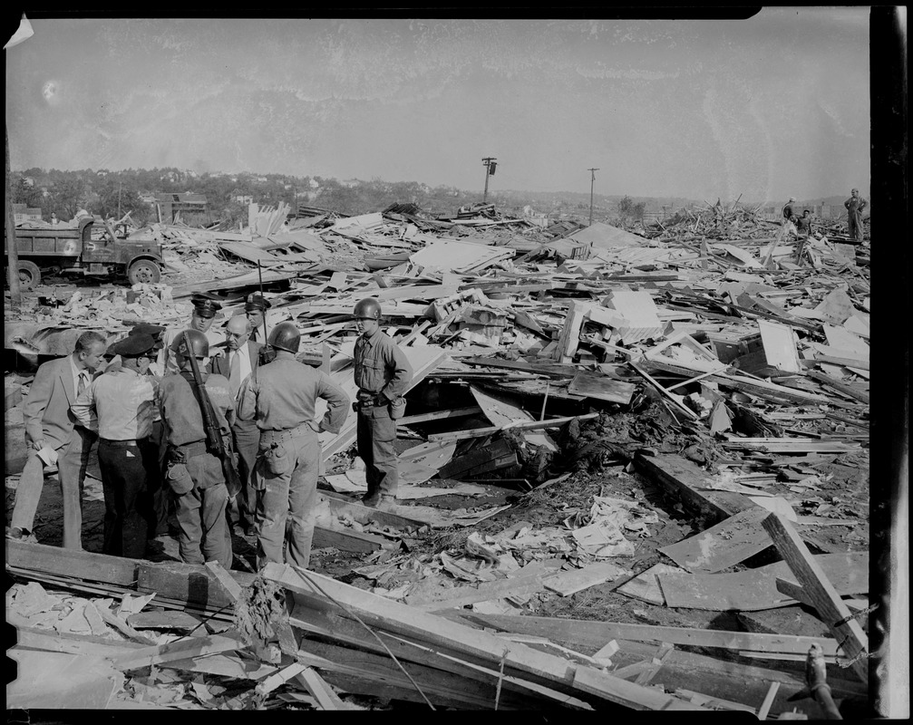 Group of men, including some in uniform, standing amid damage beside a box full of human bones