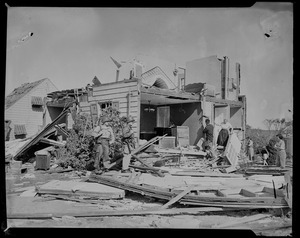 Group of people picking through house's wreckage while two men in uniform look on