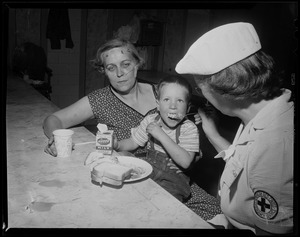 Red Cross woman feeds a boy sitting in the lap of an injured woman