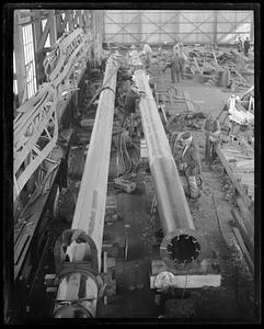 People working in a space filled with pieces of wood and two long metal tubes, probably masts for a ship