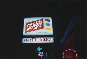 Advertisement for Schlitz beer over "Matt & Mike's" bar sign lit up at night