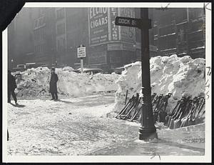 Clearing Dock Square--Hunders of workers toiled furiously today to release busy Dock square from the grip of snowdrifts. Shovels and picks of workmen are stacked against huge pile of snow at right. Narrow lanes have been cut in some directions while trucks and cars can be seen stalled in background.