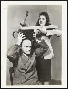The Young Lady hammers away at a nail atop a 50-pound weight on Fred Tracy's head. The performance demonstrates that a weight's resistance to motion spares Mr. Tracy's head, and proves a scientific point to student war workers.