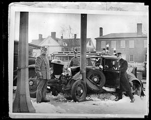 Three men in uniform stand by a wrecked car