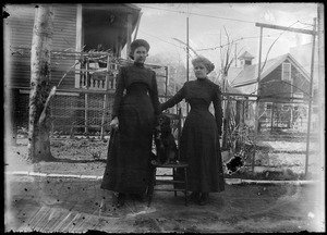 Two women standing in yard with dog on chair between them