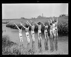 Row of naked boys standing by water, arms raised