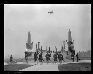 People in military uniform holding flags stand in front of John W. Weeks Bridge while an airplane files overhead