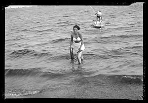 A woman stands in the water with standup paddler in background.
