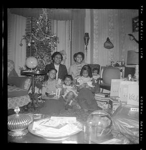 Two women and three girls are seated with a Christmas tree and a television in the background