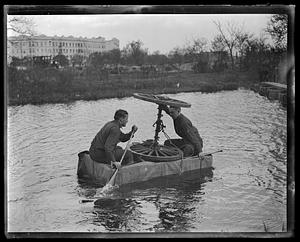 Two men on a barge with a set of wheels on an axle, one rowing